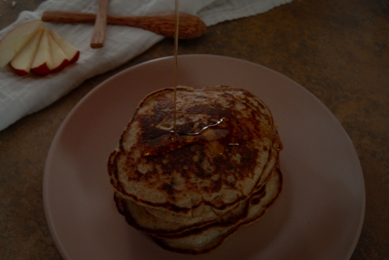 overhead shot of a stack of pancakes being covered in maple syrup. apples and wooden cutlery in the background on a grey napkin.