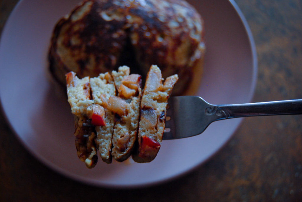 close up shot of a triangular slice of 4 apple cinnamon oat pancakes held up on a fork with the plate of pancakes in background